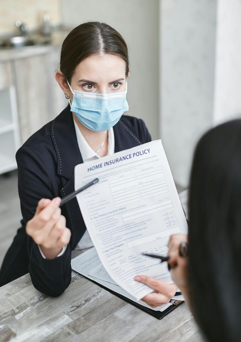 Professional woman wearing a face mask discussing a home insurance policy with a client at a desk.